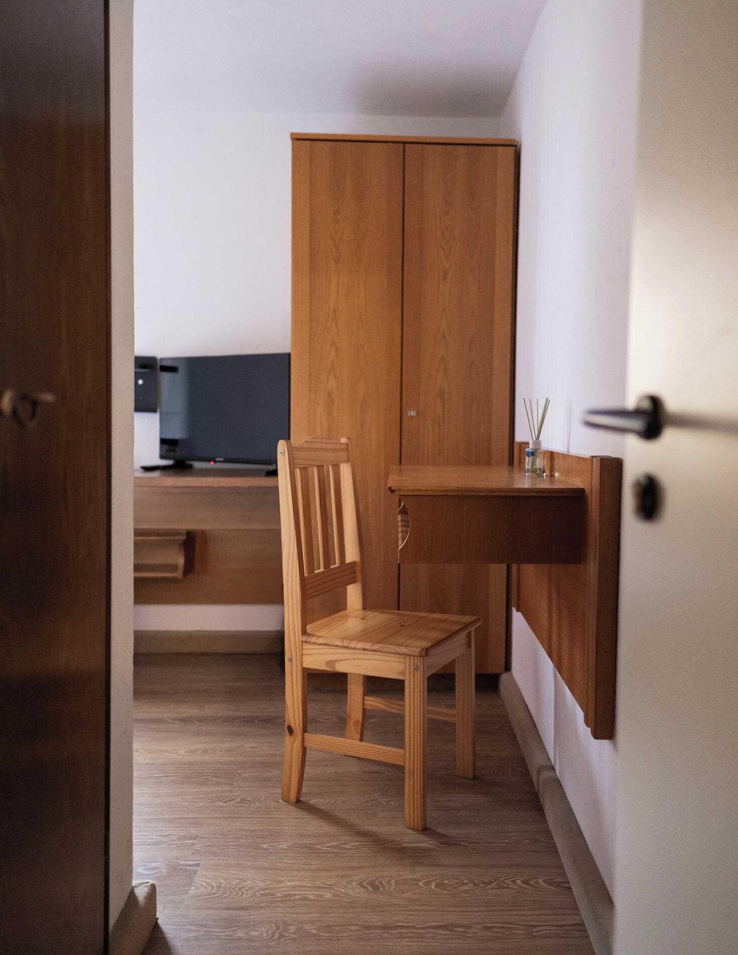 A small, wooden-framed room with a desk and chair, a wardrobe, and a television.