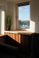 Una habitación con un asiento junto a la ventana con vistas a un lago, una planta en maceta y un banco de madera.