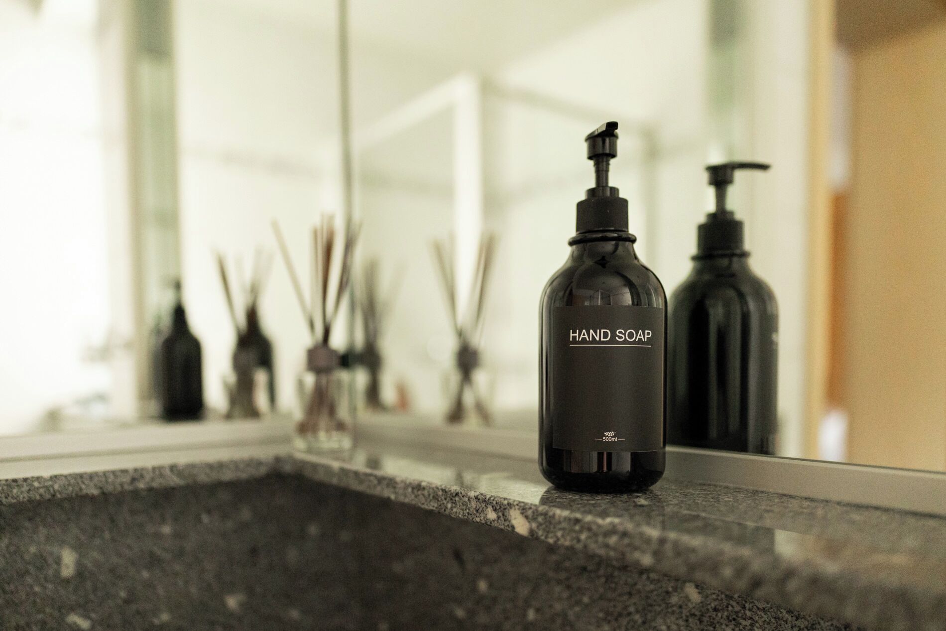 A hand soap bottle on a countertop with a mirror reflecting various bathroom items.
