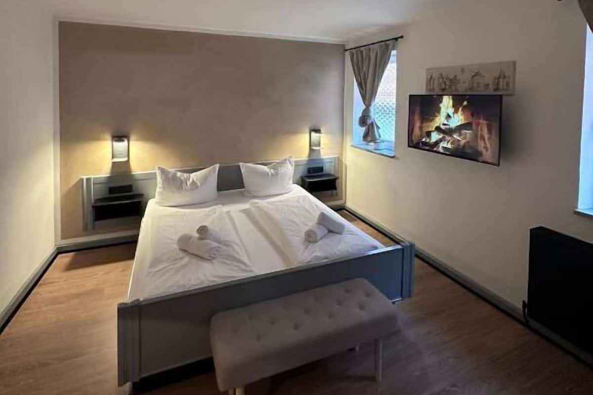 Une chambre à coucher moderne avec un grand lit, des tables de chevet, un banc et une image de cheminée sur le mur.
