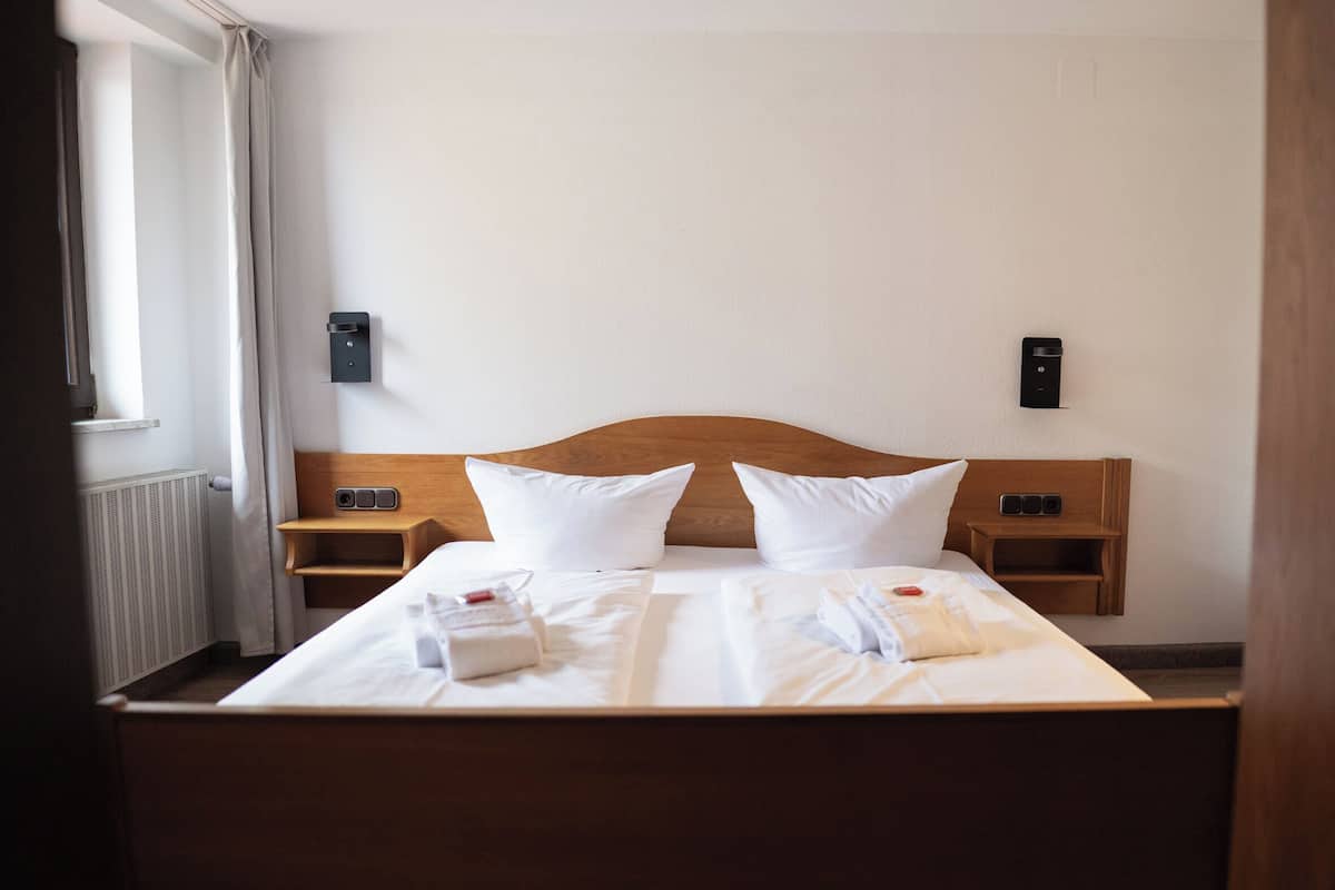 Un lit bien fait, avec du linge de lit blanc, deux oreillers et une tête de lit en bois. Il y a deux tables de chevet avec des lampes et un thermostat fixé au mur.