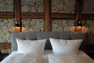 Cama con ropa blanca y dos almohadas, frente a pared de piedra con vigas de madera.