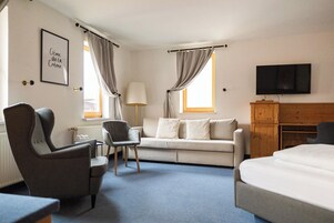 Habitación de hotel con cama, dos sillones, sofá, escritorio, lámpara, televisor y cortinas.