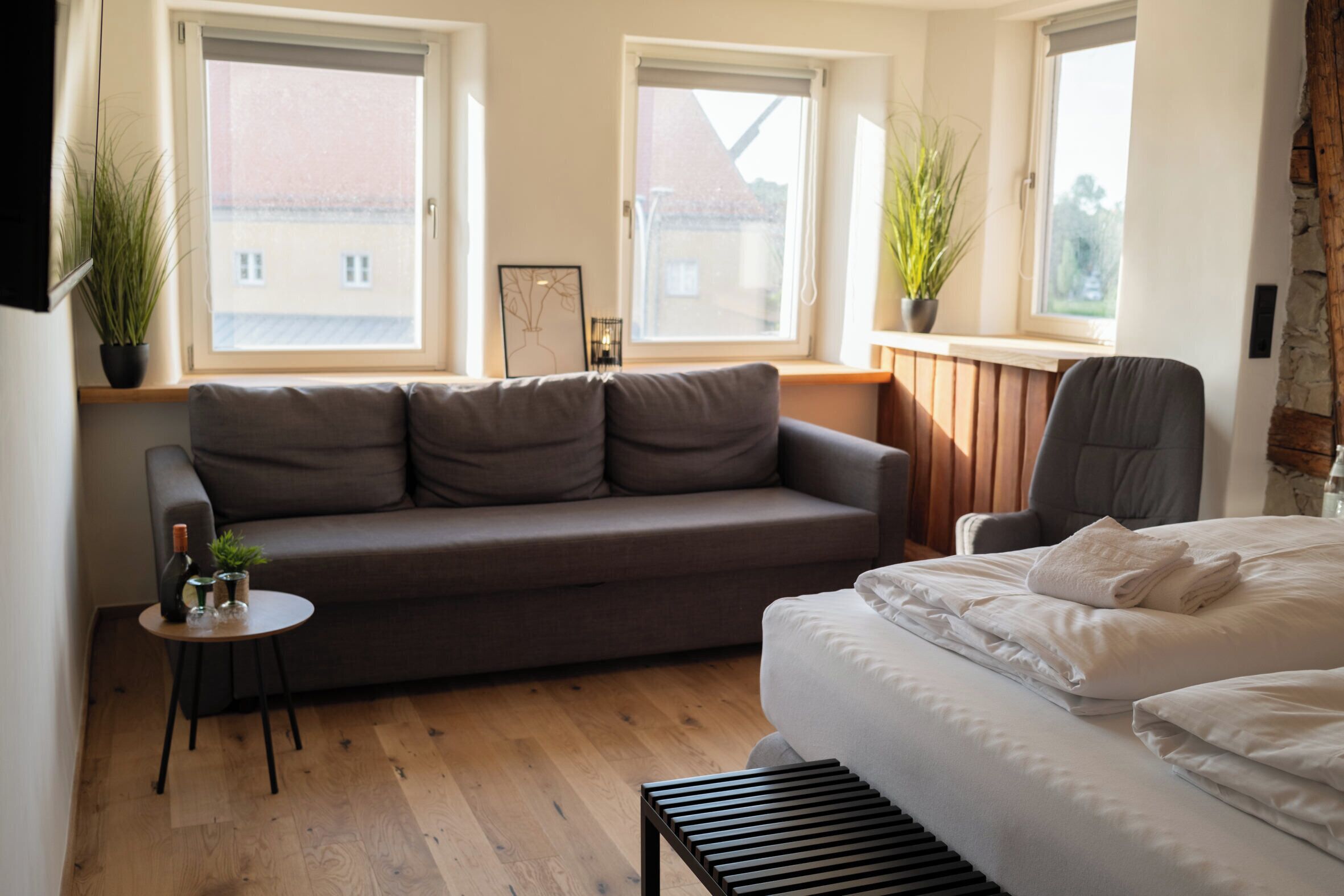 Un dormitorio moderno con un sofá gris, una mesita redonda pequeña, una cama con ropa blanca y dos ventanas con plantas en macetas.