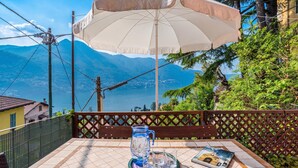 A table with a pitcher, glasses, and a magazine, set under a large umbrella overlooking a lake and mountains. - Casa Valleggia (Moltrasio)