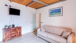 A room with a wooden ceiling, a flat-screen TV mounted on the wall, a wooden cabinet with decorative items, a beige sofa, and a framed landscape painting. - Casa Valleggia (Moltrasio)