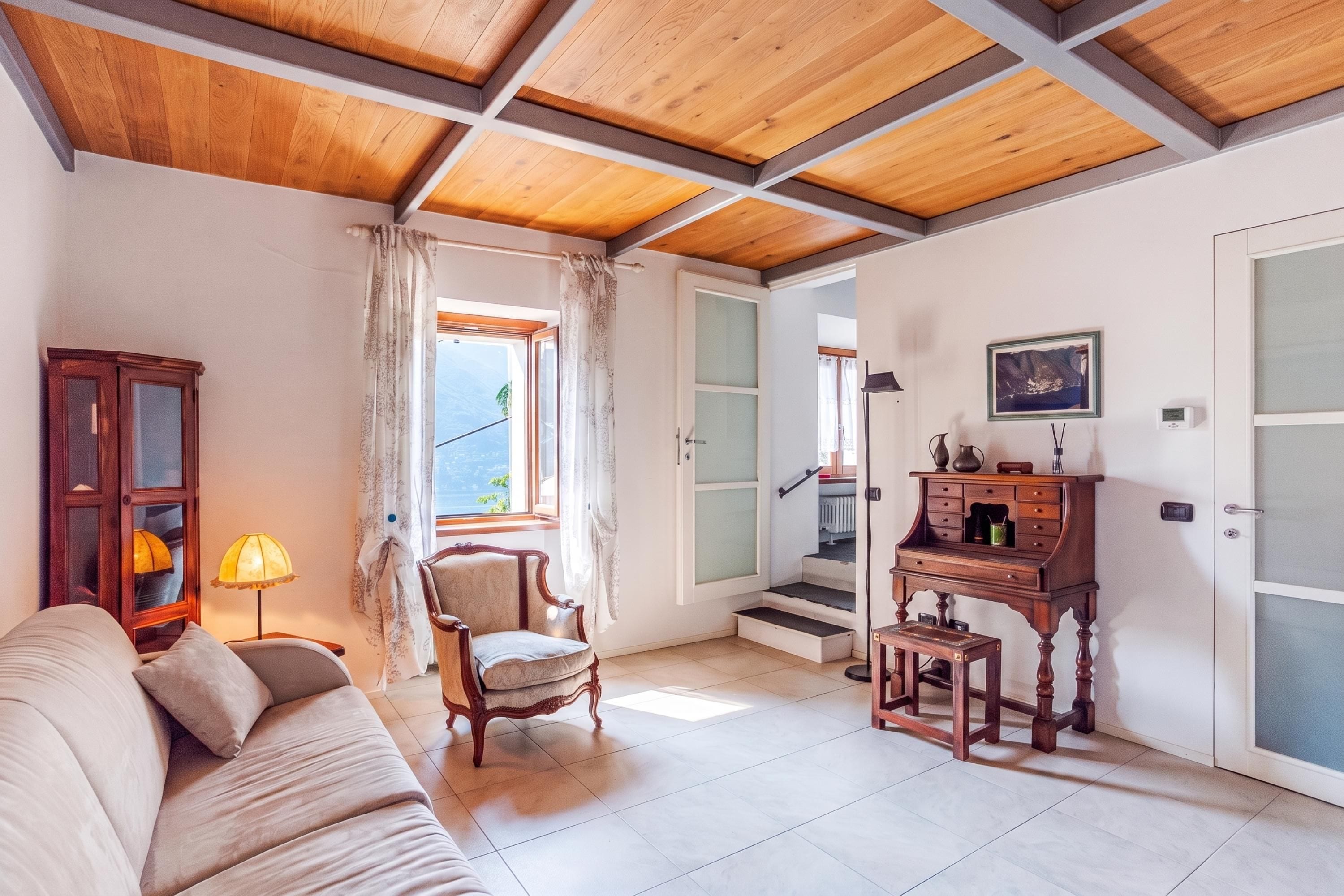 Una habitación con techo de madera, un sofá, una silla, un escritorio con cajones y una ventana con cortinas.
