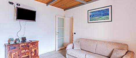 Una habitación con techo de madera, un televisor de pantalla plana en la pared, un mueble de madera con adornos, un sofá color beige y un cuadro con un paisaje enmarcado.