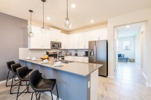 Una cocina moderna con armarios blancos, una isla grande con taburetes de bar, electrodomésticos de acero inoxidable e iluminación colgante.