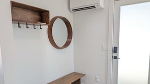 Un pasillo con un banco de madera, un estante de madera con ganchos, un espejo redondo y una unidad de aire acondicionado.