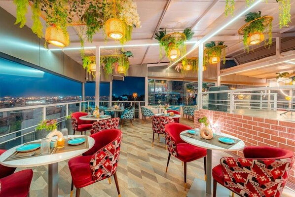 A rooftop restaurant with red chairs, tables, hanging plants, and a city view.