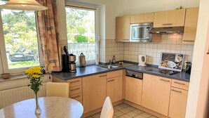 A modern kitchen with light wood cabinets, a stainless steel microwave, and a dishwasher. There is a window with curtains, a vase with flowers on the table, and a tiled floor. - Vacation home Maria by Interhome (Zinnowitz)