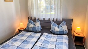 A double bed with a checkered blue and white bedspread, two matching pillows, a bedside table with a lamp, and a window with sheer curtains. - Vacation home Maria by Interhome (Zinnowitz)
