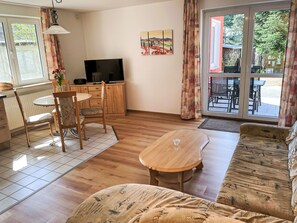 Ein Wohnzimmer mit Sofa, Couchtisch, Fernseher, Esstisch mit Stühlen und einem Fenster mit Vorhängen.