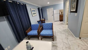 Una sala de estar con una mesa de centro blanca, dos sofás azules estampados, un dispensador de agua grande y un control remoto.