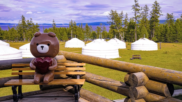 Una escultura de oso sosteniendo un corazón en un banco de madera, con yurtas y un paisaje boscoso al fondo.
