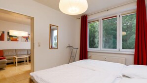 Ein Schlafzimmer mit einem großen Bett, einem Fenster mit roten Vorhängen, einem Heizkörper und Blick auf Bäume.