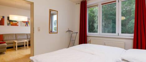 A bedroom with a large bed, a window with red curtains, a radiator, and a view of trees outside.