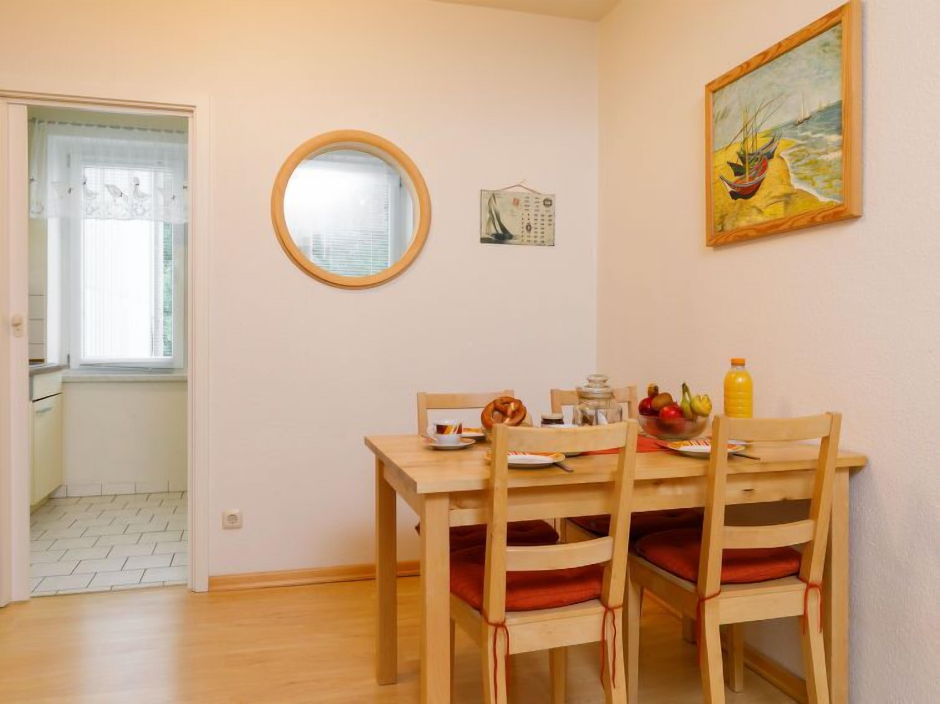 Un coin repas avec une table et des chaises en bois, un miroir rond, un tableau et une fenêtre avec des stores.