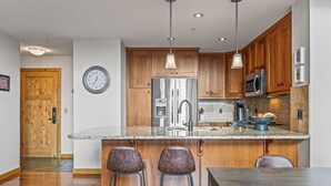 A modern kitchen with wooden cabinets, stainless steel appliances, and a central island with bar stools. - Wake Up to Three Sisters | Penthouse Condo (Canmore)