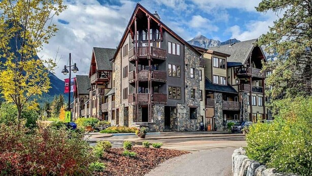 A multi-story lodge with stone and wood exteriors, balconies, and a mountain backdrop. - Wake Up to Three Sisters | Penthouse Condo (Canmore)