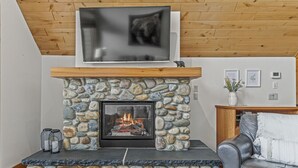 A stone fireplace with a TV above it, a stone bench, and a grey sofa in a living room with wooden ceiling beams. - Wake Up to Three Sisters | Penthouse Condo (Canmore)