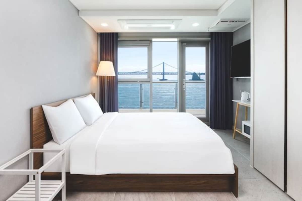 A modern hotel room with a bed, a desk, a chair, a TV, and a view of a bridge over water.