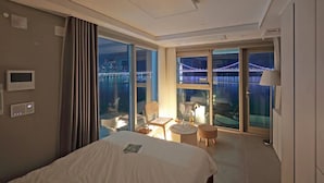 A modern hotel room with a large bed, a seating area with a table and chairs, and a view of a cityscape through large windows.