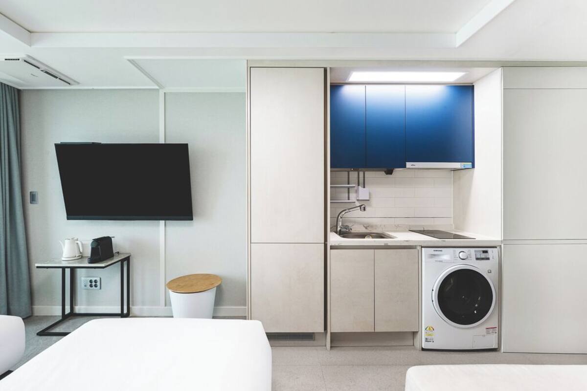 Une chambre d’hôtel compacte comprenant un lit, une petite table avec une bouilloire et une tasse, un lave-linge, un évier et un téléviseur fixé au mur.