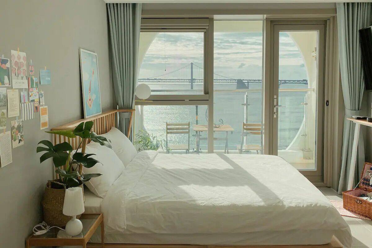 Une chambre à coucher avec un lit, une table de chevet, une lampe et une plante en pot. Il y a un balcon avec vue sur la mer et un pont.