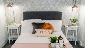 Un dormitorio ordenado con cama, dos mesitas de noche, una mesilla auxiliar, un cojín decorativo, una bandeja con una planta y tazas, y un papel tapiz estampado.