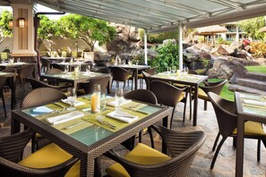 Un área de comedor al aire libre con sillas y mesas de mimbre, con rocas y follaje de fondo.