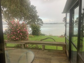 A wooden deck with a glass table, overlooking a lake with a pink flowering tree and a grassy area. - Waterfront - 3 bedroom, 2 bathroom, peaceful, private with- sunny conservatory (Glenbrook)