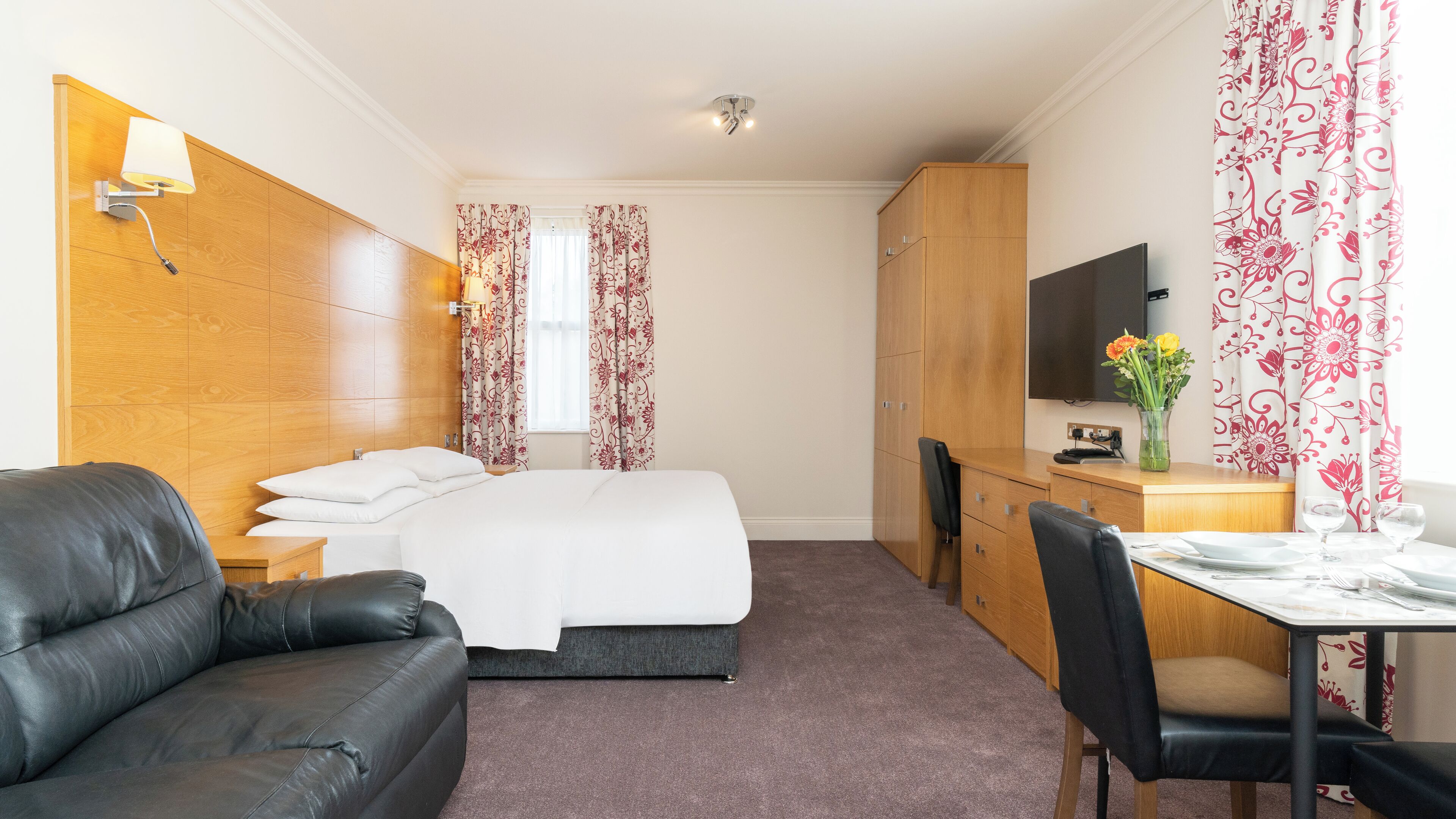 A hotel room with a bed, a desk, a TV, a leather sofa, and a dining table with chairs.