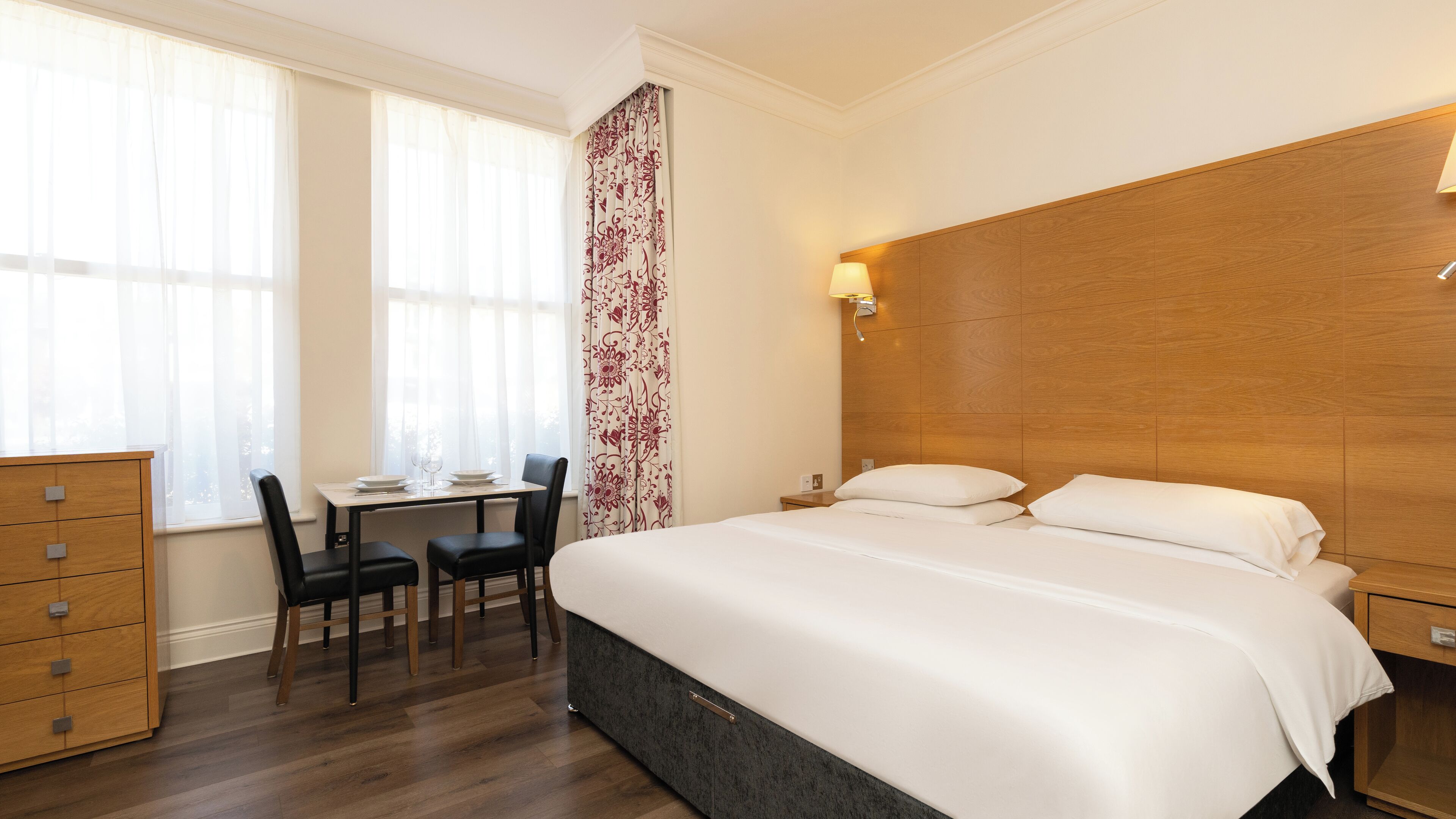Una camera d'albergo con un grande letto, una cassettiera in legno, una scrivania con sedie e una finestra con tende trasparenti.