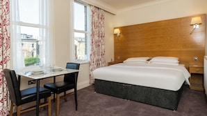 Een hotelkamer met een bed, nachtkastje, lamp, raam met gordijnen en een tafel met stoelen.
