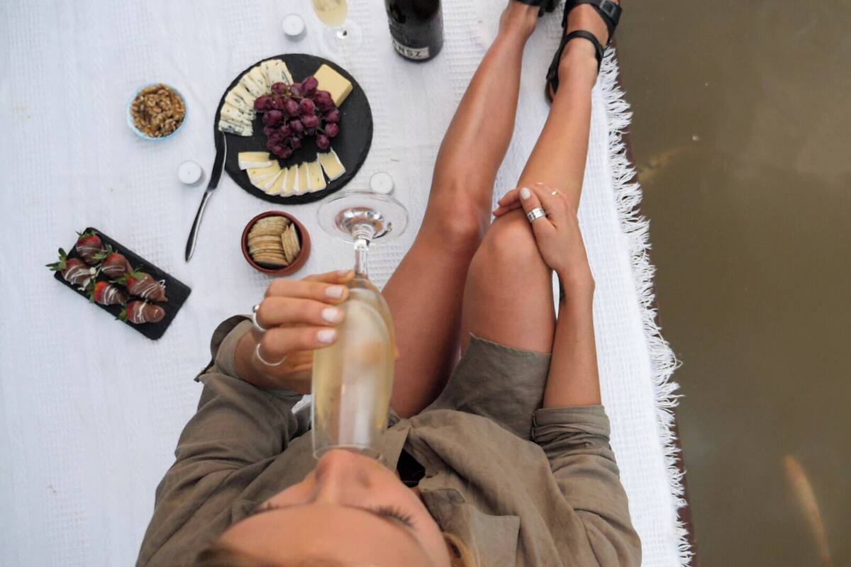 A person lying on a white towel with a bottle of white wine, a plate of cheese and grapes, and a bowl of nuts.