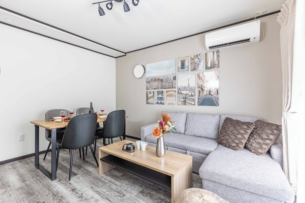A modern living room with a grey sofa, a wooden coffee table, and a dining area with chairs and a table set for two.