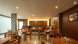 A hotel restaurant with wooden furniture, a central dining area, and a view of the kitchen in the background. - Hotel The King's (Port Blair)