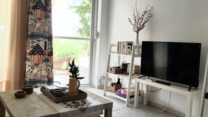 Ein Wohnzimmer mit großem Flachbildfernseher, Bücherregal, einem Fenster mit Vorhängen und einem kleinen Tisch mit Dekoration.