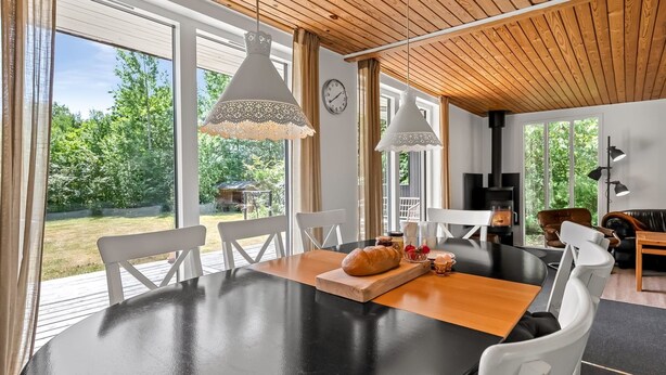Un espace repas moderne avec une table ronde, des chaises blanches et un banc en bois. On y trouve des luminaires suspendus, une horloge et une vue sur l’extérieur.