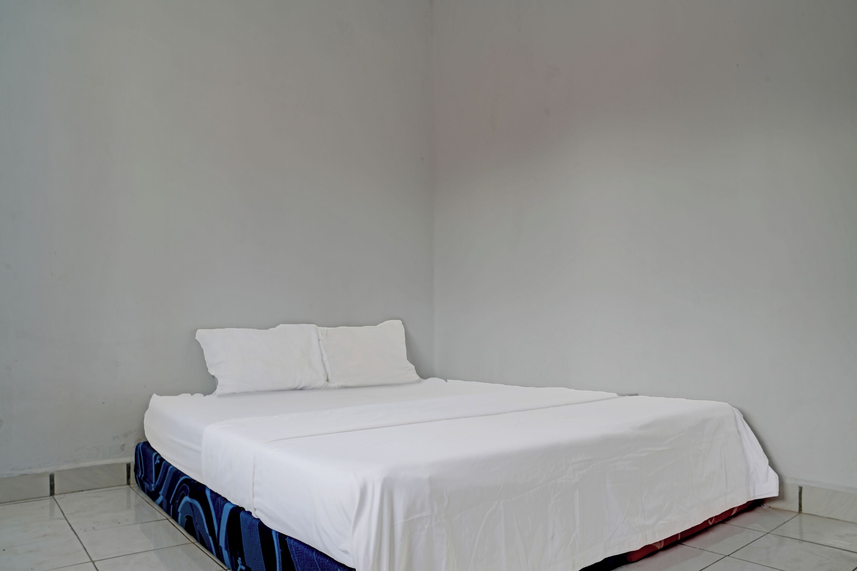 Habitació amb un llit individual, roba de llit blanca, una falda amb dibuixos blaus i vermells, i parets blanques.