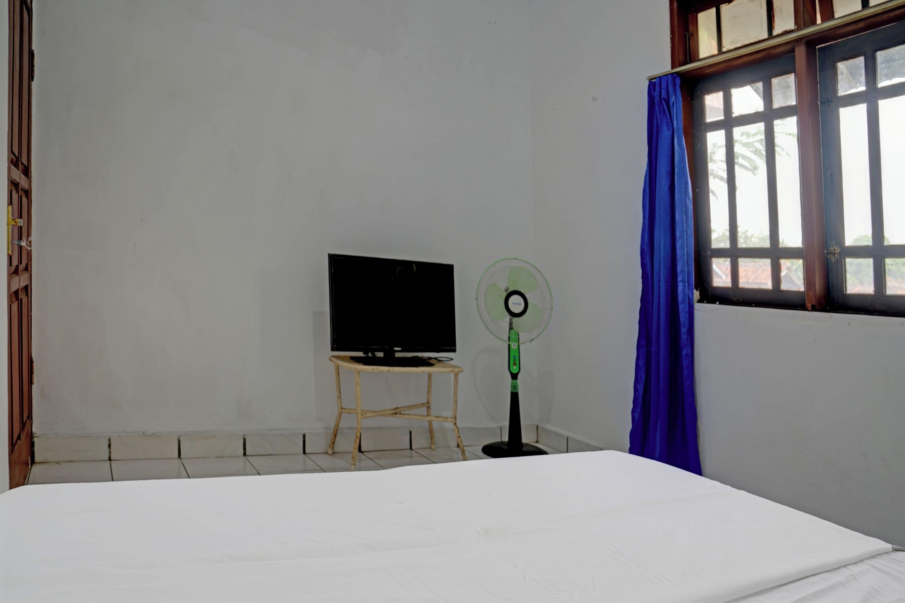 A room with a bed, a television on a small table, a standing fan, and a window with a blue curtain.