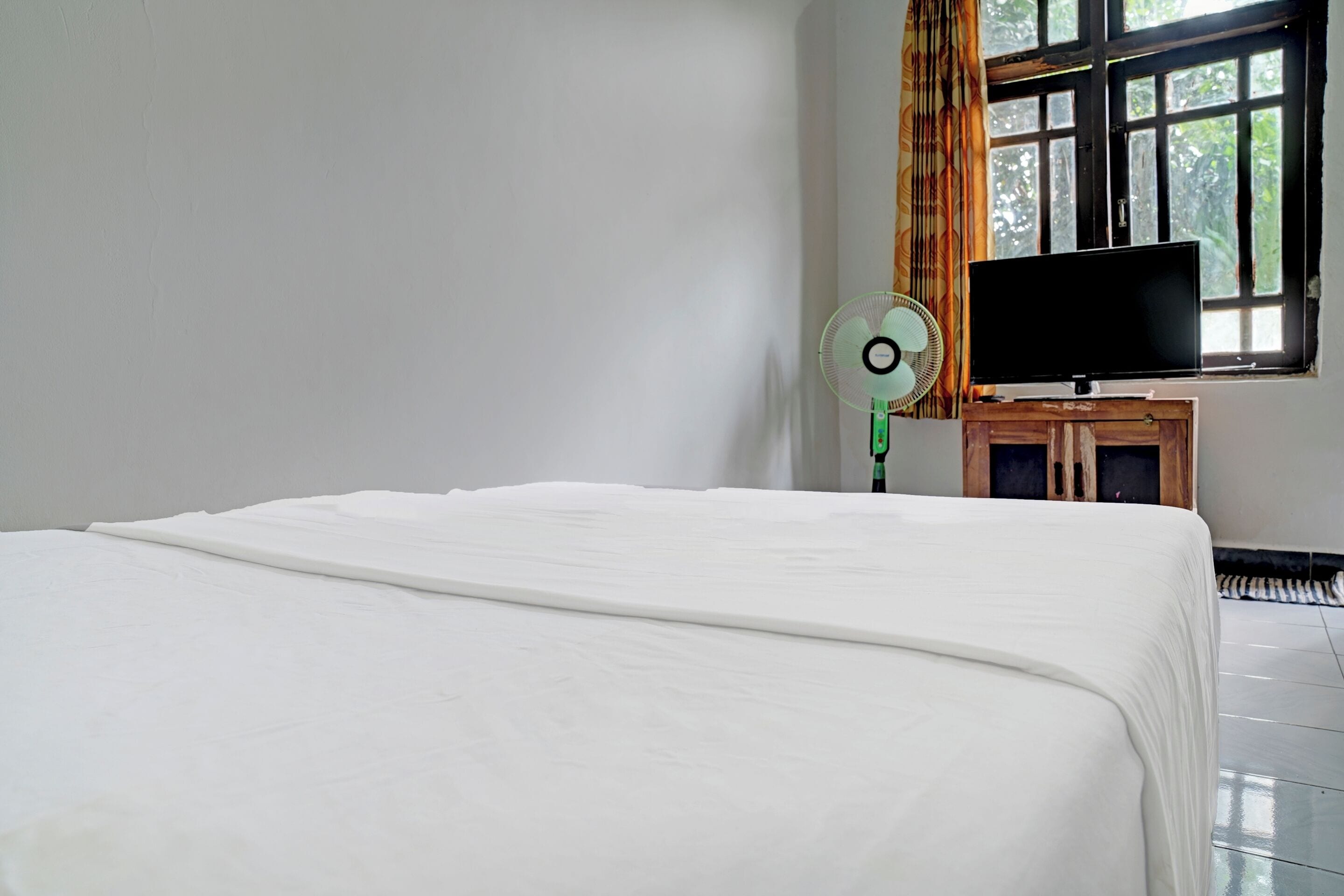 Une chambre à coucher avec un lit, un téléviseur posé sur un meuble en bois, un ventilateur et une fenêtre avec des rideaux.