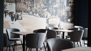 Un café con sillas y mesas modernas, vista a una calle histórica con coches antiguos y edificios.