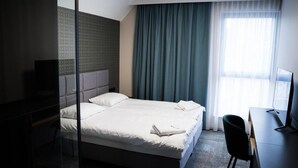 Una habitación de hotel con cama, escritorio, silla, televisor y ventana con cortinas.
