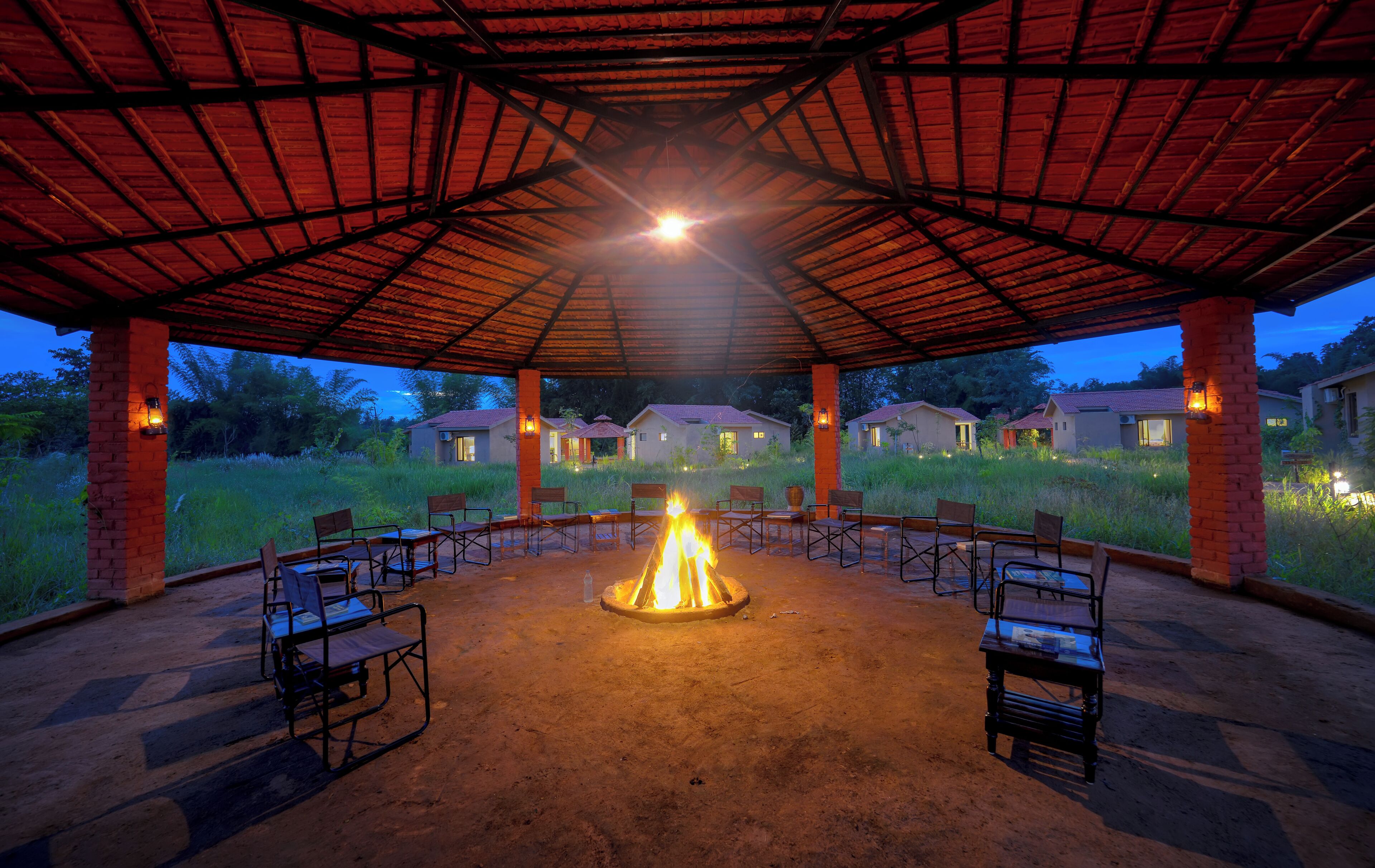An outdoor pavilion with a central fire feature, surrounded by chairs and tables, set against a backdrop of residential buildings.