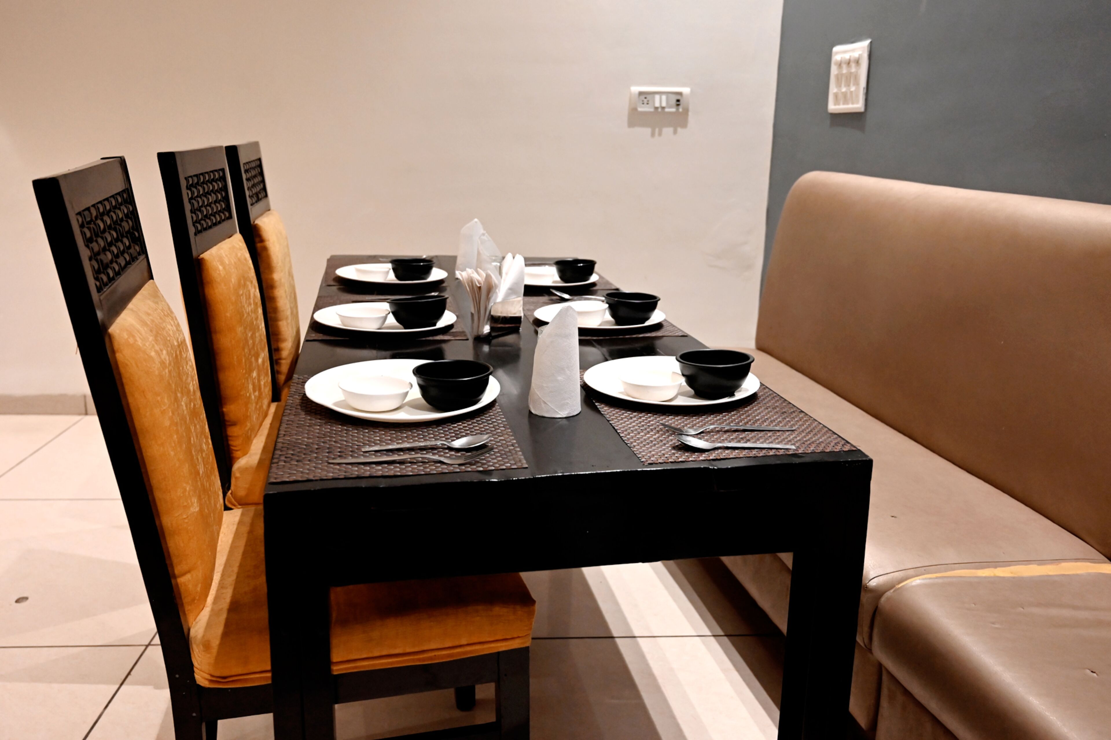 A dining table set with black bowls, white plates, and cutlery, accompanied by a brown upholstered bench and a wall socket.