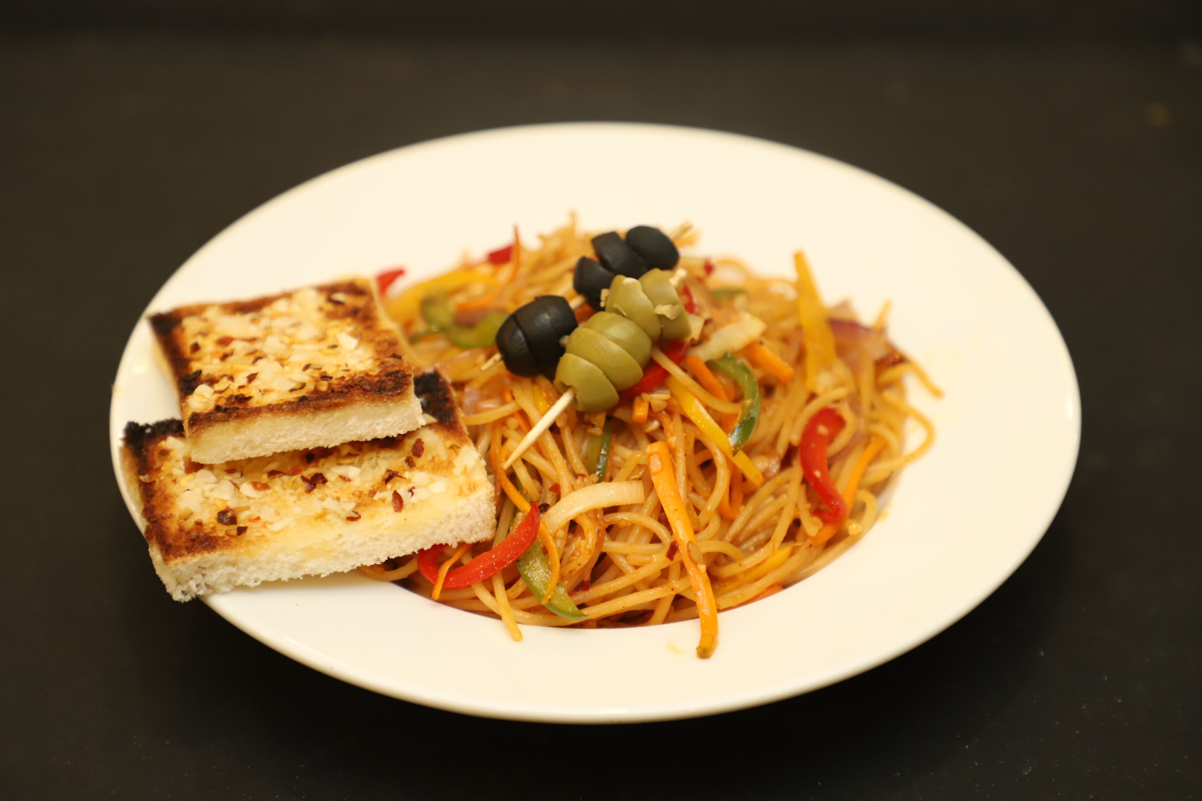 Un piatto di noodles con verdure e olive, servito con due fette di pane grigliato.