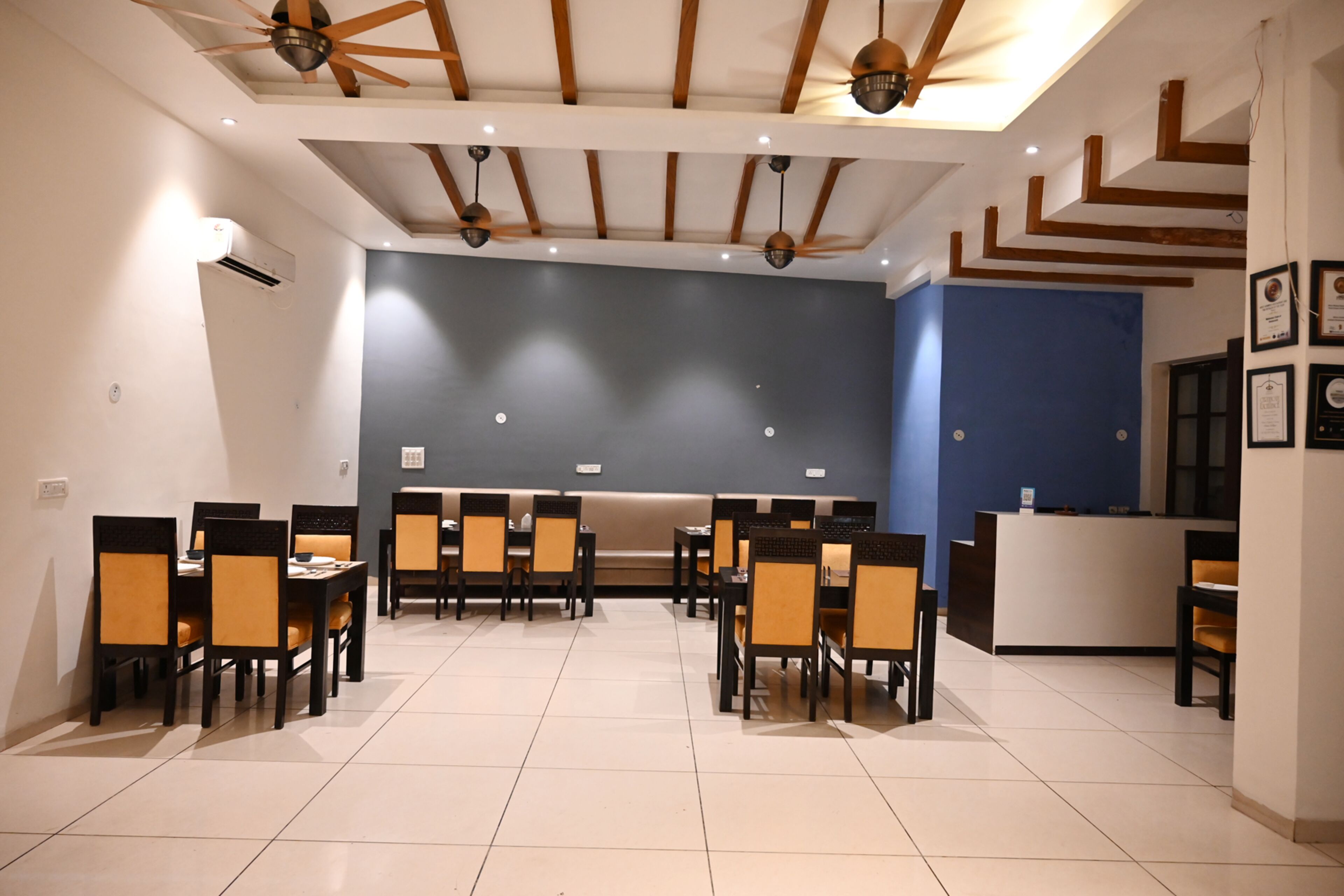 A modern dining area with multiple tables and chairs, a reception desk, and a ceiling with wooden beams and pendant lights.